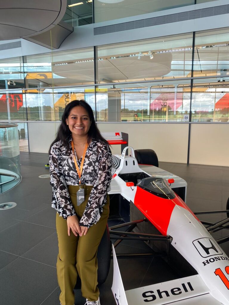 Jess smiles and leans on Aryton Senna's McLaren at McLaren HQ.