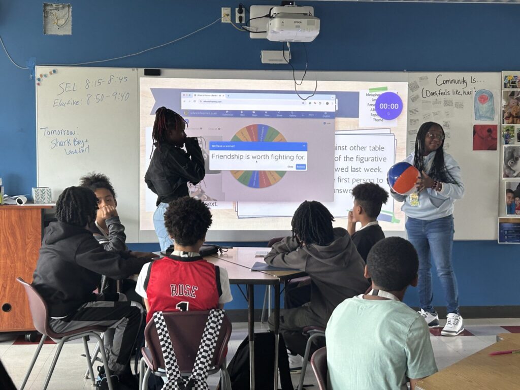 Students in a classroom, playing a game involving passing a ball. A screen, reading "Friendship is Worth Fighting For" is in the background. 